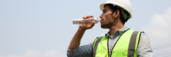 A man in a hard hat and reflector vest drinks a bottle of water on a sunny day.